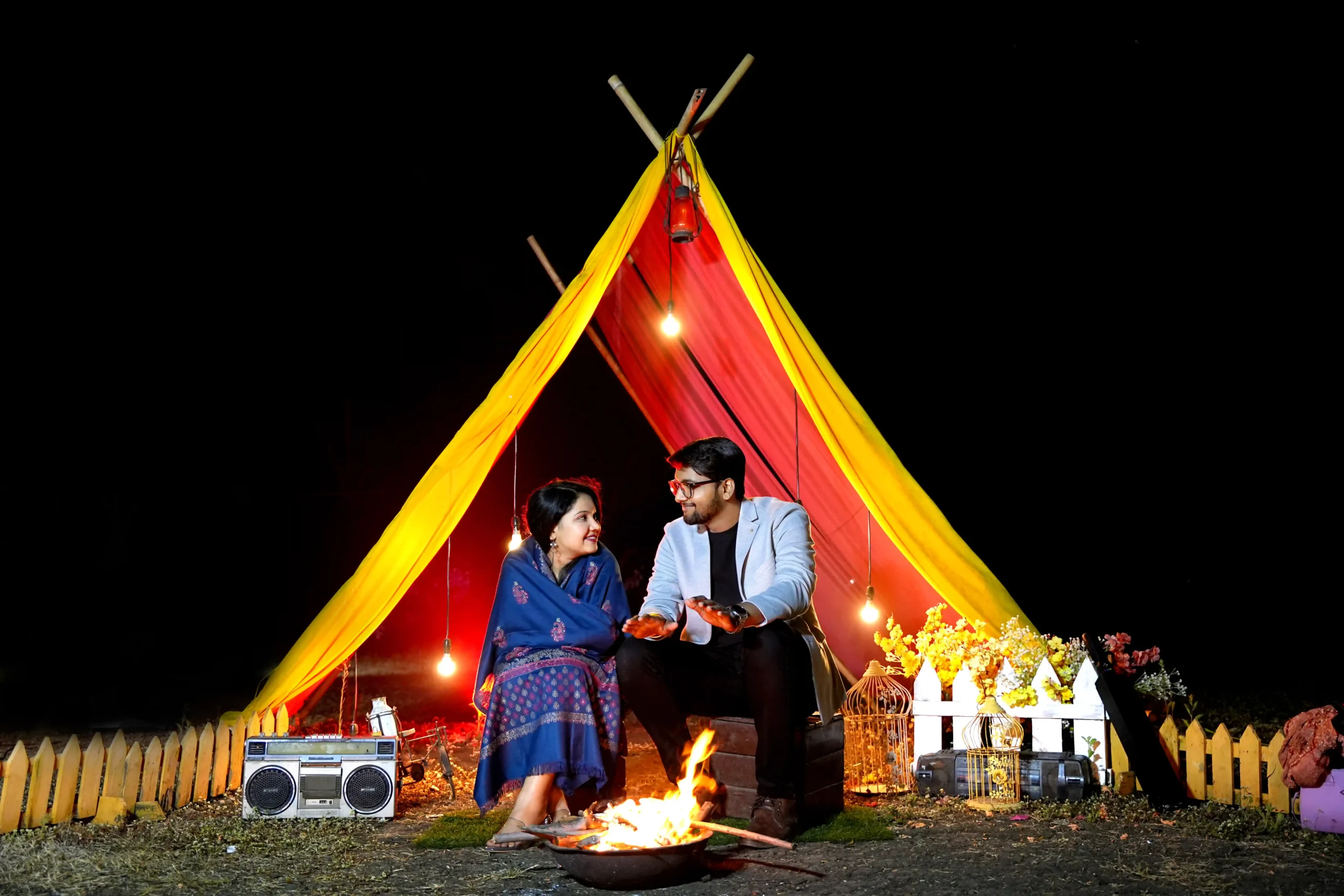 Couple sitting by a campfire during a night pre-wedding shoot inside a decorated teepee tent with warm lights and props – Capturing Vivah (The CV Studios).